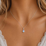 A close-up of a woman's neck and chest wearing a delicate silver chain necklace with a teardrop-shaped iridescent natural moonstone pendant.