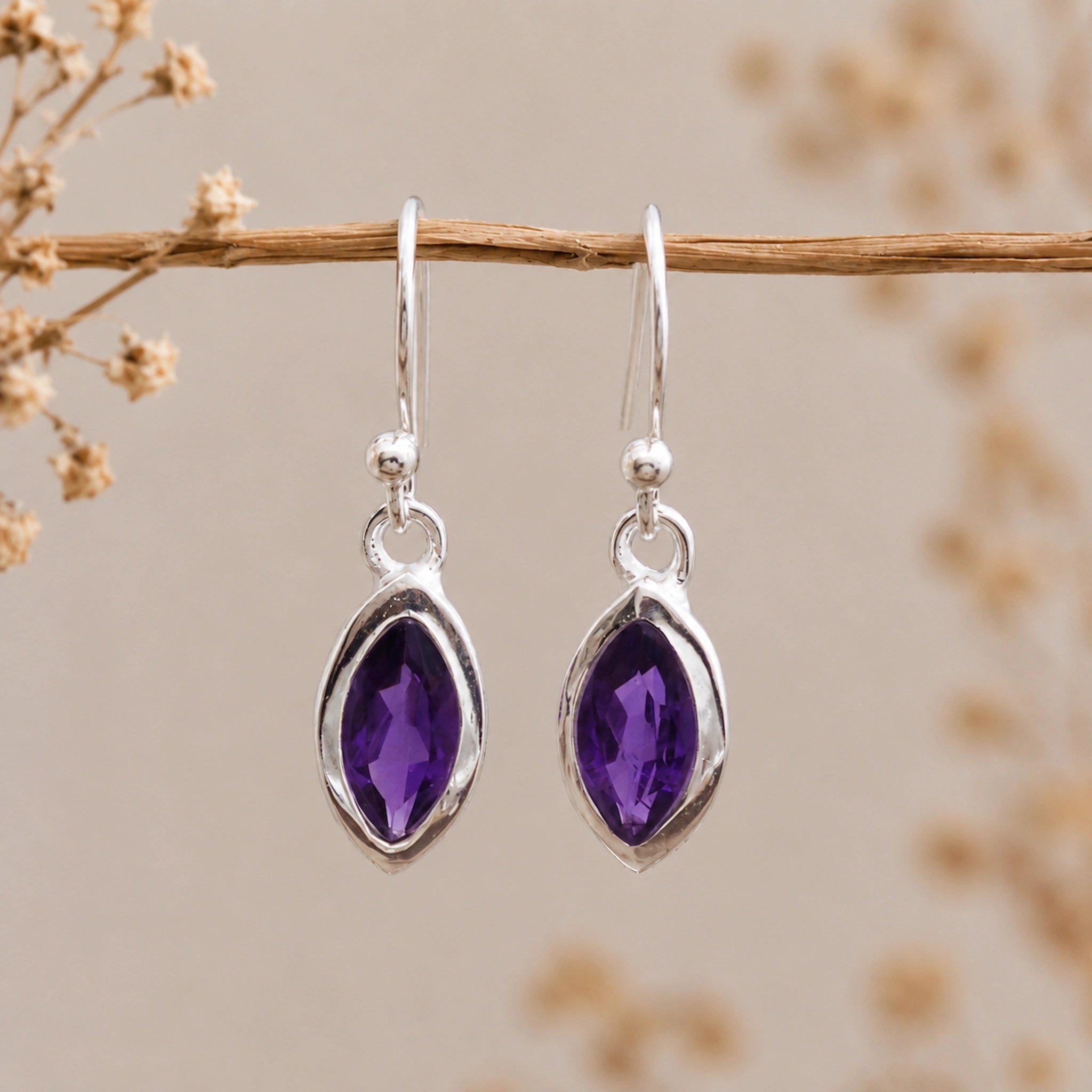 Silver earrings with marquise shaped purple amethyst gemstones on a branch with dried flowers.