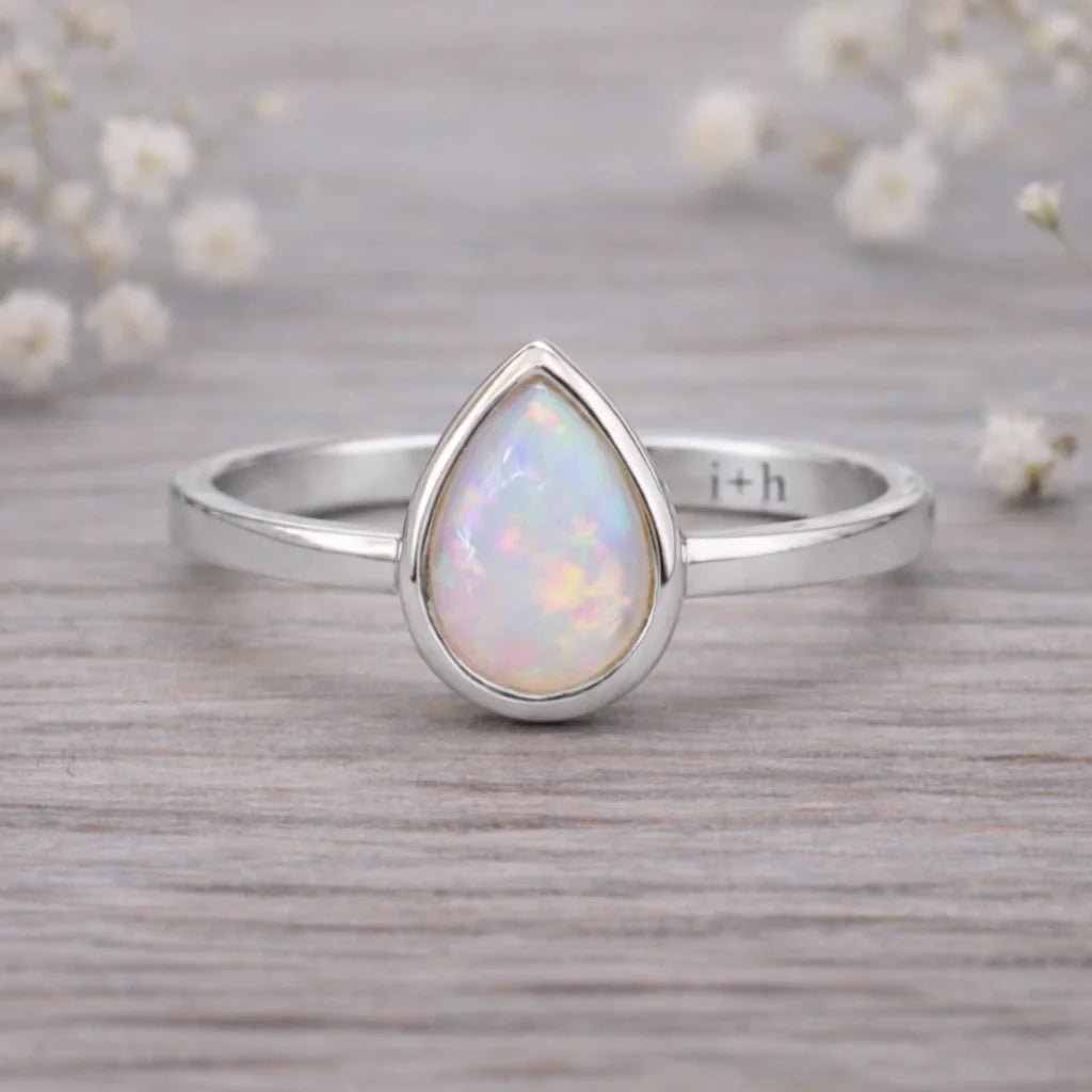 Silver ring with a teardrop-shaped opal stone on a wooden surface with flowers in the background