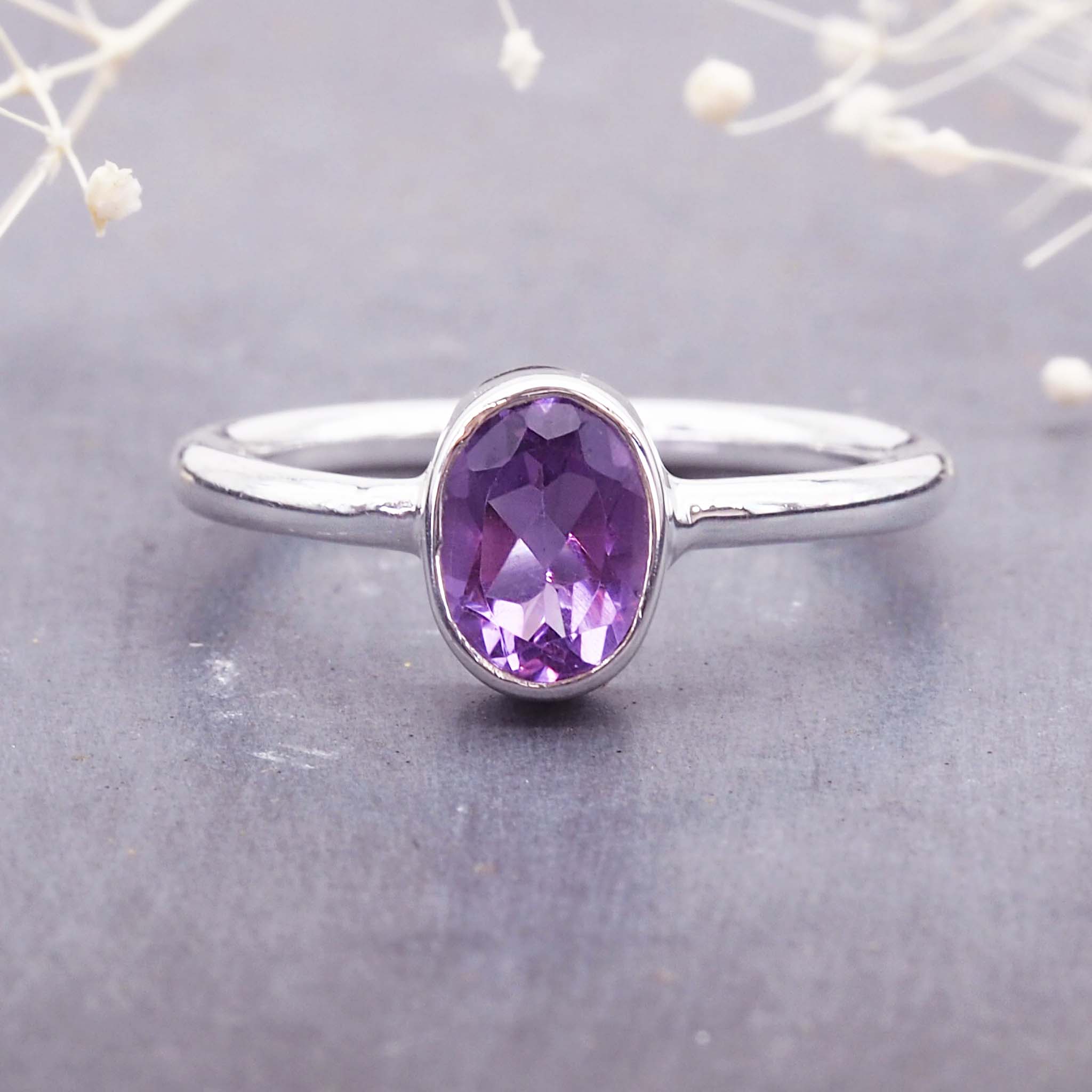 Sterling Silver ring with a natural amethyst gemstone on a grey surface with blurred purple flowers in the background