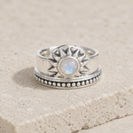 A sterling silver sunburst ring with a central round iridescent moonstone, resting on a textured beige stone surface.