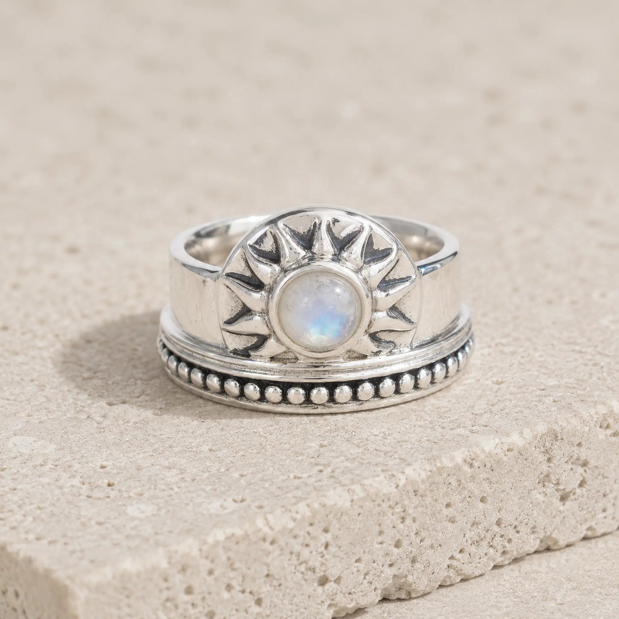 A sterling silver sunburst ring with a central round iridescent moonstone, resting on a textured beige stone surface.