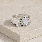 A sterling silver ring with a sunburst design and a central round iridescent moonstone, resting on a textured beige stone block.