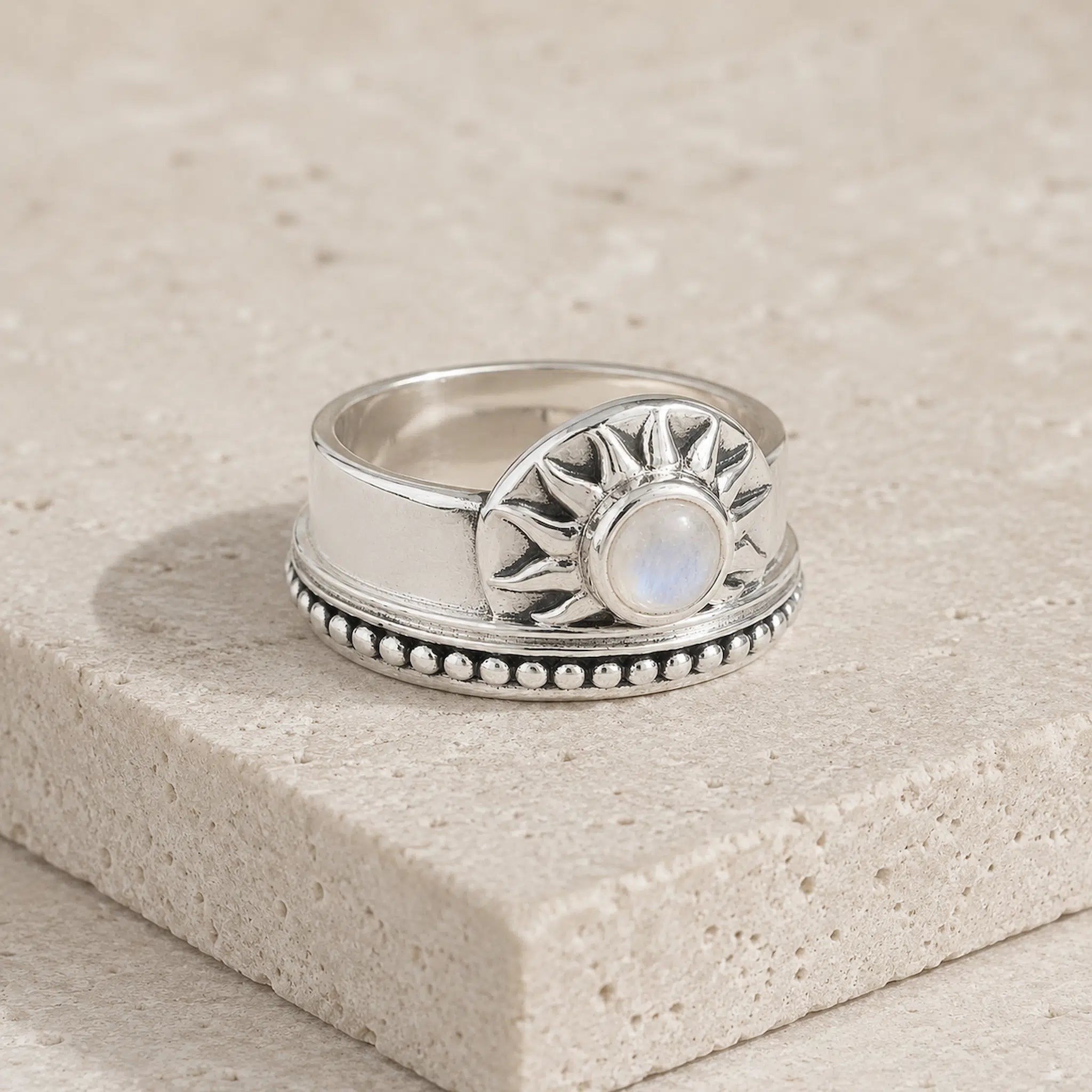 A sterling silver ring with a sunburst design and a central round iridescent moonstone, resting on a textured beige stone block.