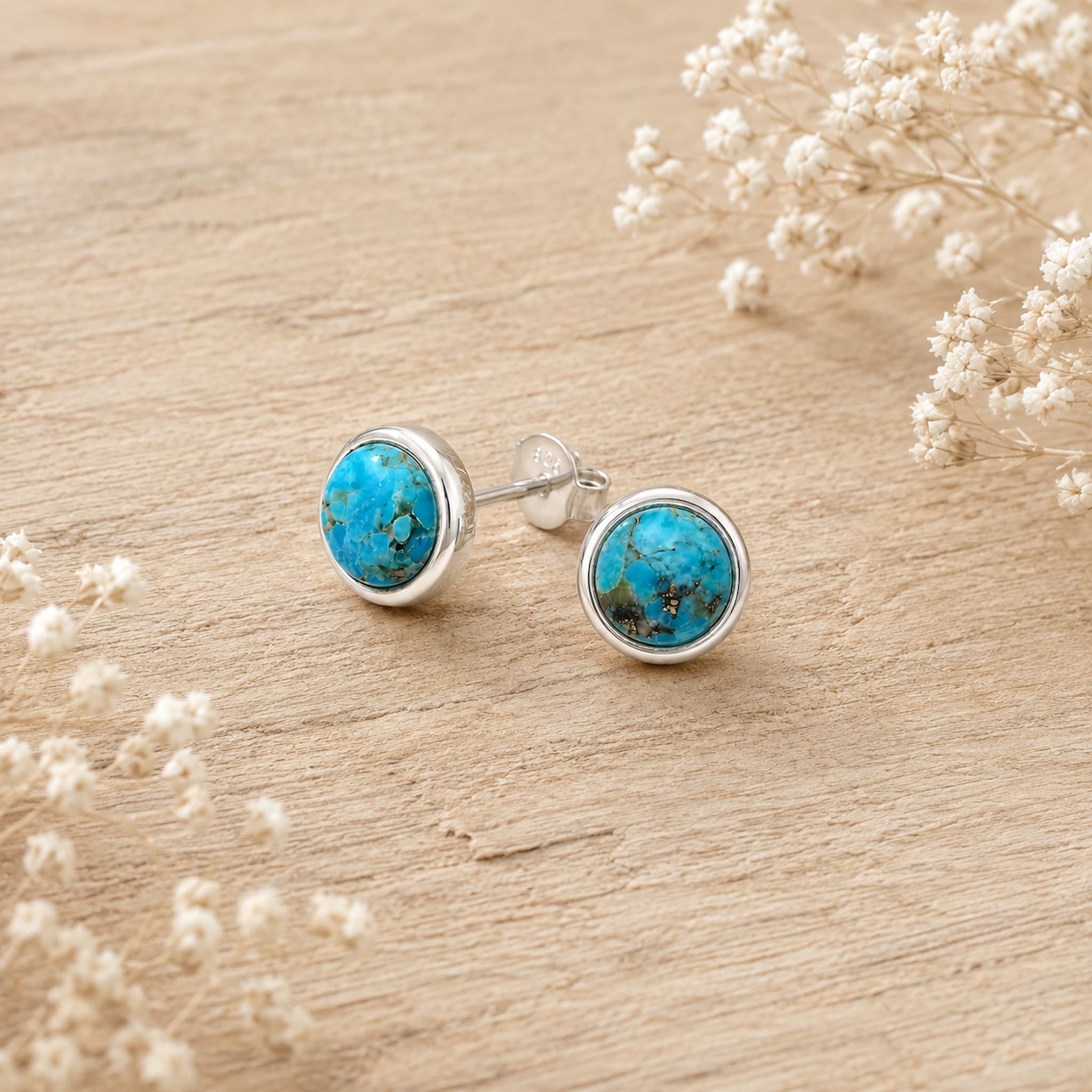 A pair of round stud earrings with vibrant turquoise-blue stone centers and silver bezels, displayed on a light wood surface with dried white flowers.