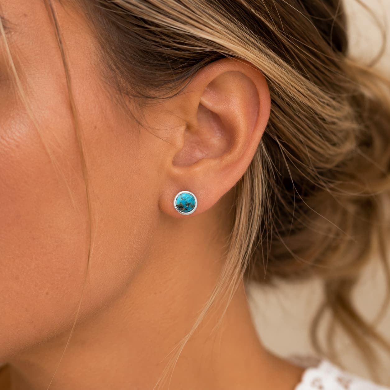 A close-up of a woman's ear wearing a small round stud earring with a bright turquoise-blue stone in a silver-toned bezel setting.