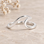 A polished silver wave-shaped ring with a flat band, resting on a textured beige stone surface with dried baby's breath flowers in the background.