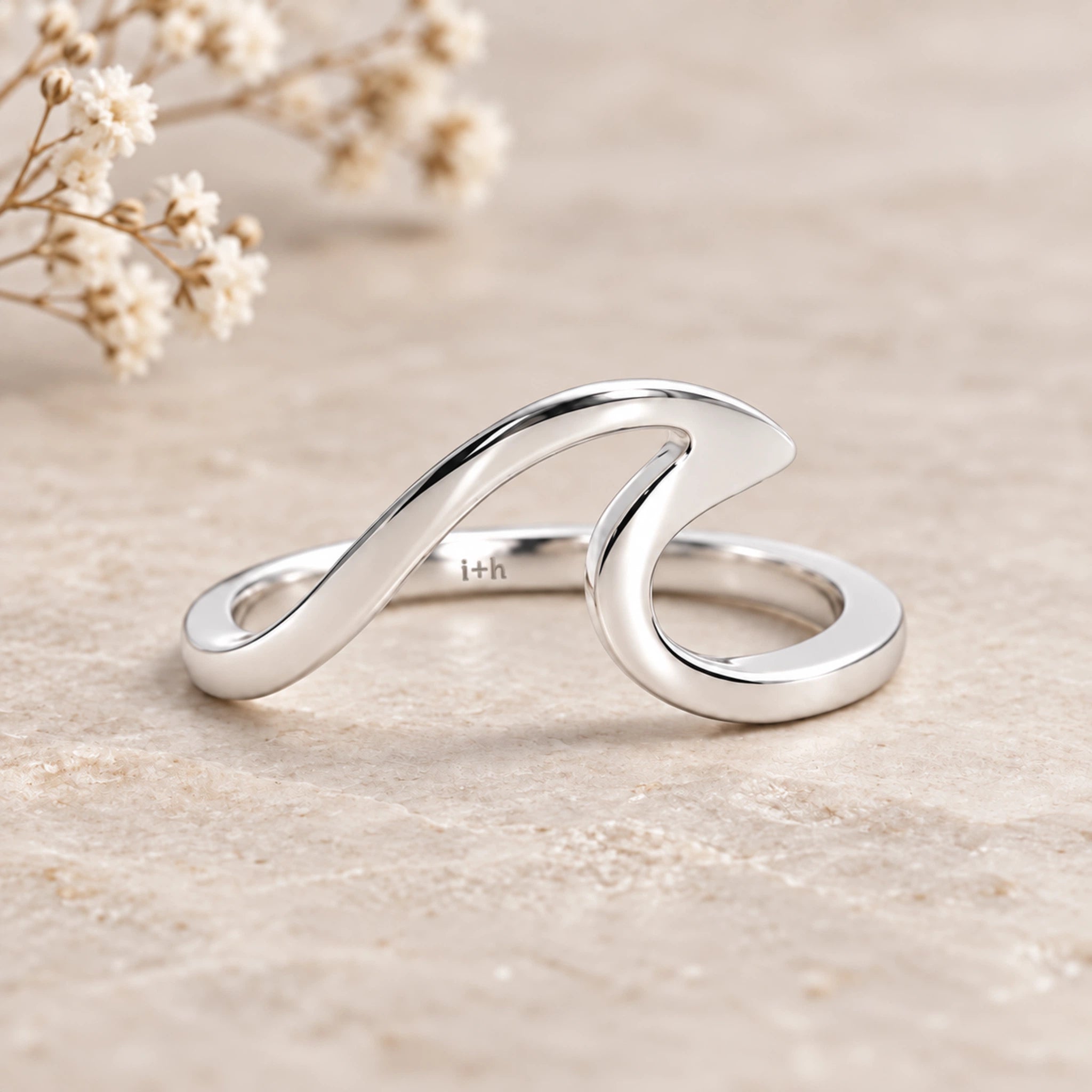 A polished silver wave-shaped ring with a flat band, resting on a textured beige stone surface with dried baby's breath flowers in the background.