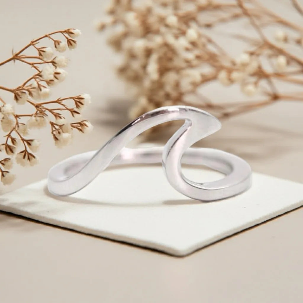 Silver ring with a wavy design on a white surface with beige flowers in the background