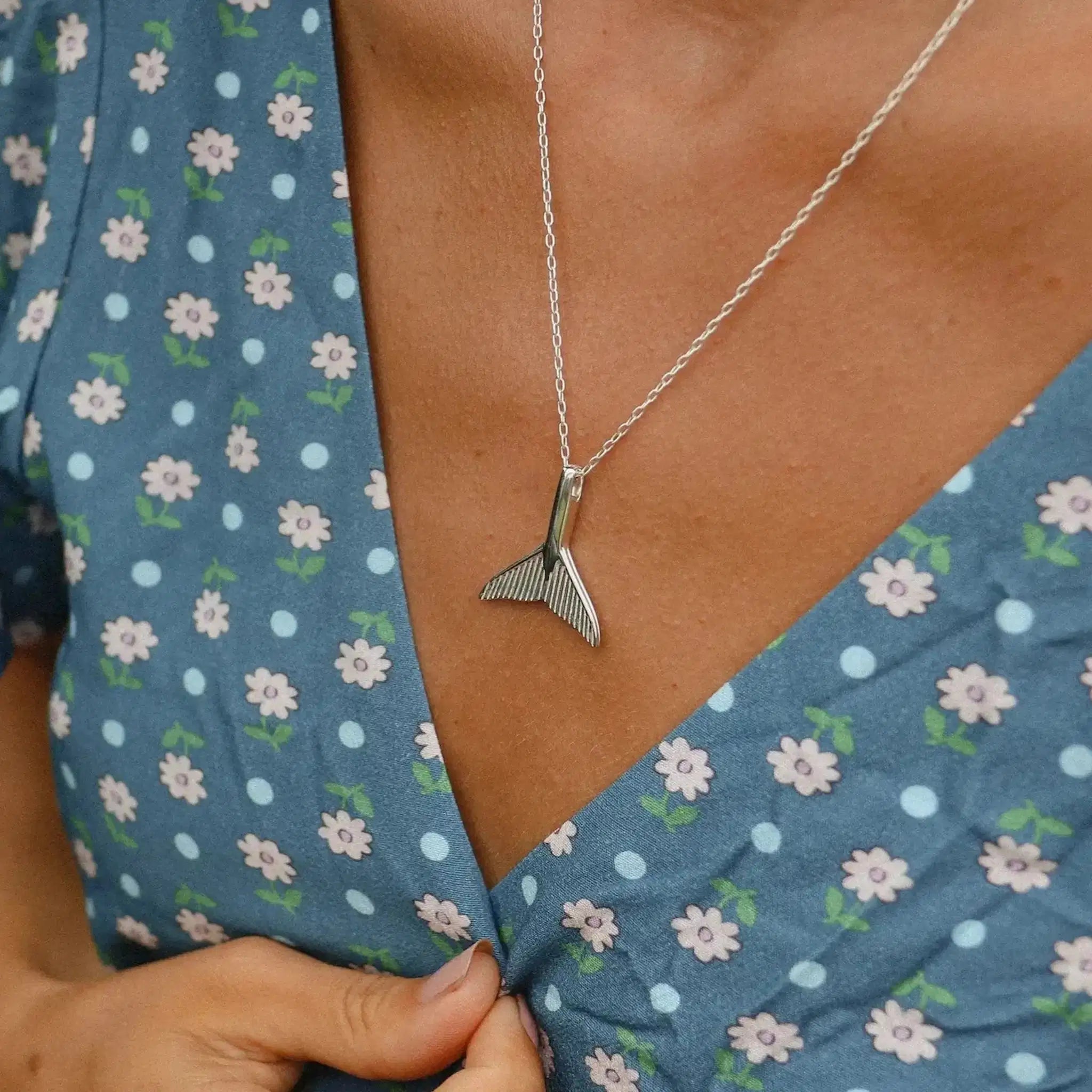 Sterling silver whale tail pendant necklace worn on a model, resting flat against a floral-patterned blue dress.