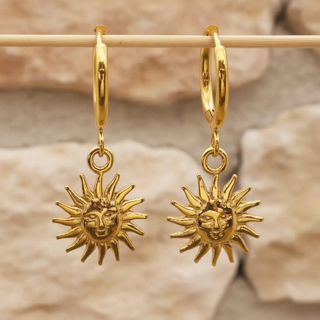 Sunburst charm gold huggie earrings in waterproof stainless steel hanging from a wooden rod
