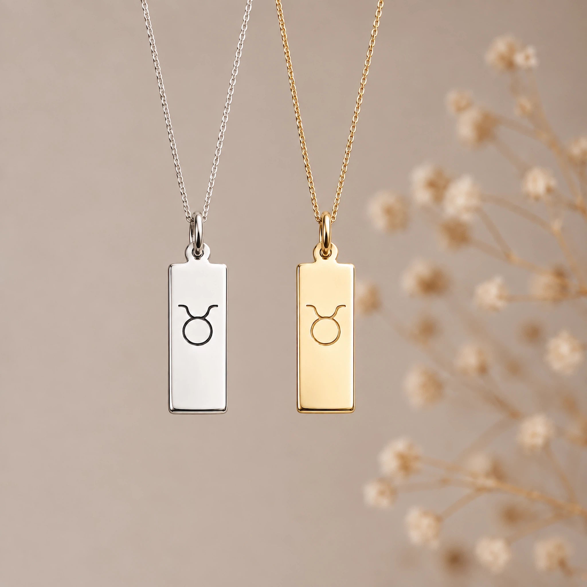 Two taurus necklaces with rectangular pendants hanging against a soft beige background, one in silver and one in gold, both engraved with a taurus zodiac line glyph symbol. Dry flowers are blurred in the background on the right.