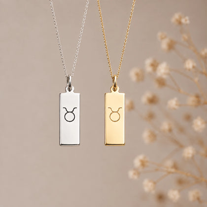 Two taurus necklaces with rectangular pendants hanging against a soft beige background, one in silver and one in gold, both engraved with a taurus zodiac line glyph symbol. Dry flowers are blurred in the background on the right.