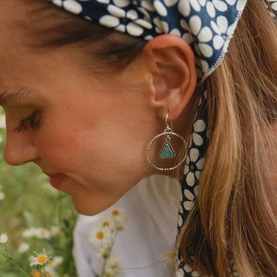 woman wearing sterling silver earrings with a hoop that has a triangular turquoise stone hanging in the middle in a field of daisies