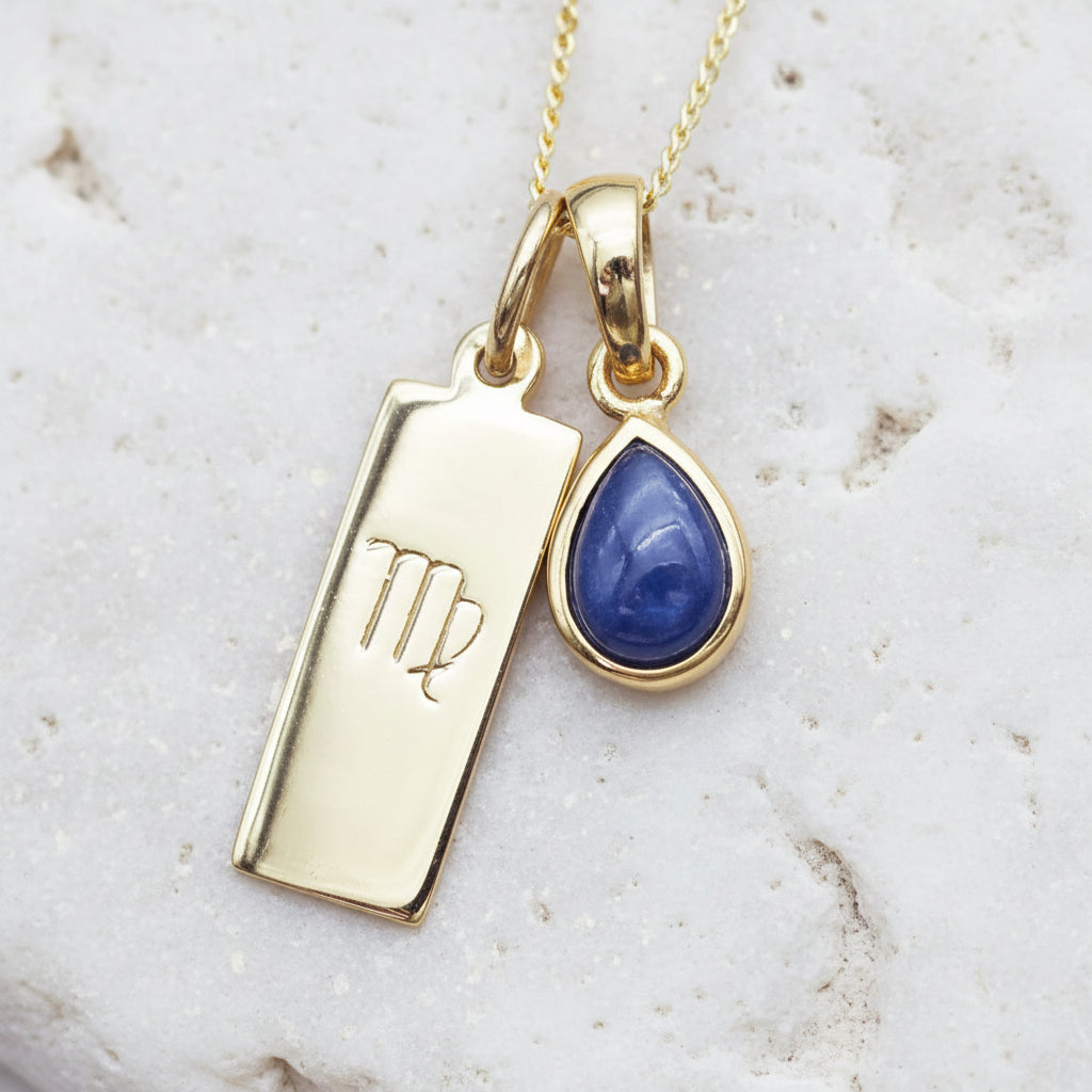 Gold necklace with Virgo zodiac sign pendant and blue teardrop natural sapphire pendant on a textured background
