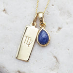 Gold necklace with Virgo zodiac sign pendant and blue teardrop natural sapphire pendant on a textured background