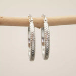 Silver hoop earrings with textured design hanging on a wooden stick against a beige background