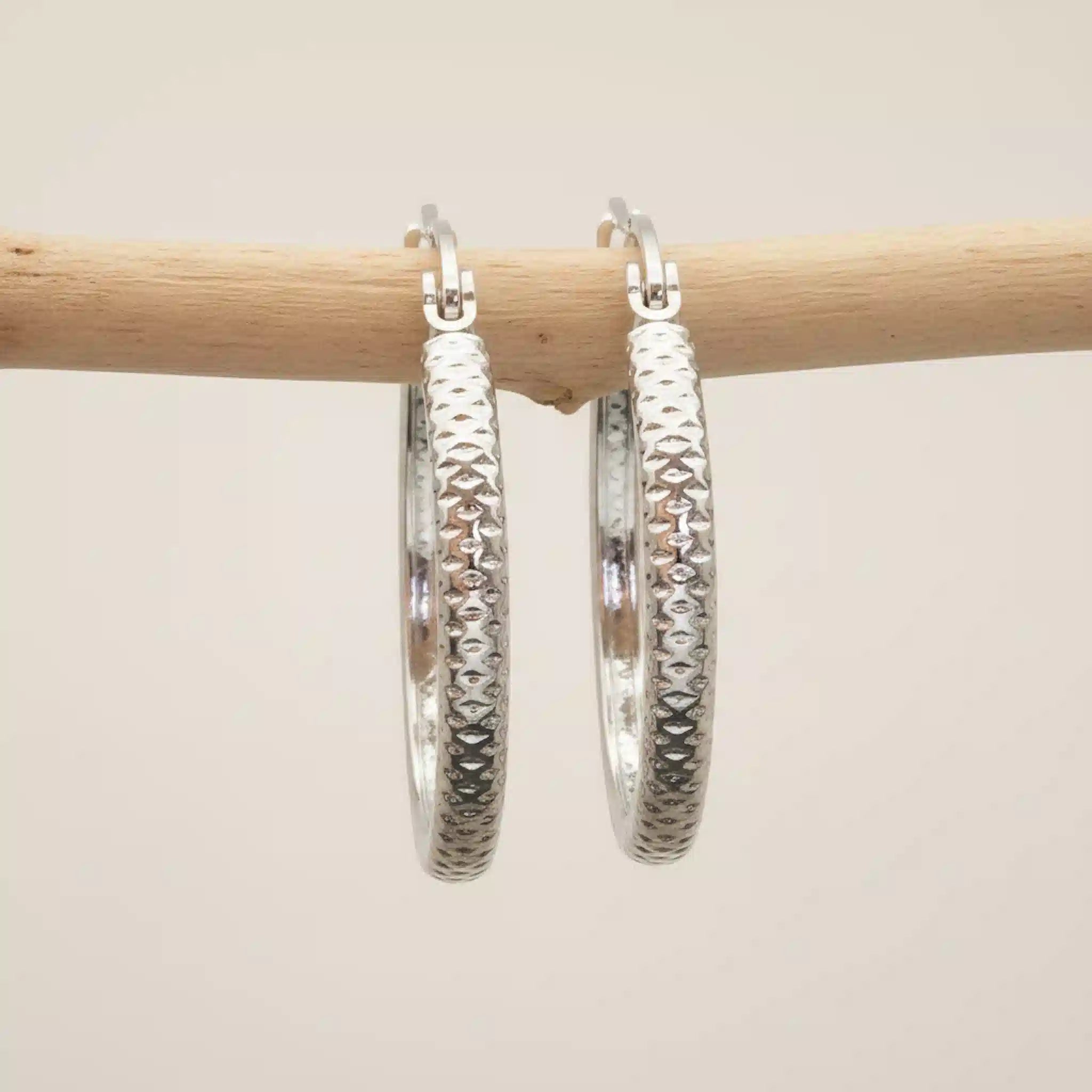 Silver hoop earrings with textured design hanging on a wooden stick against a beige background