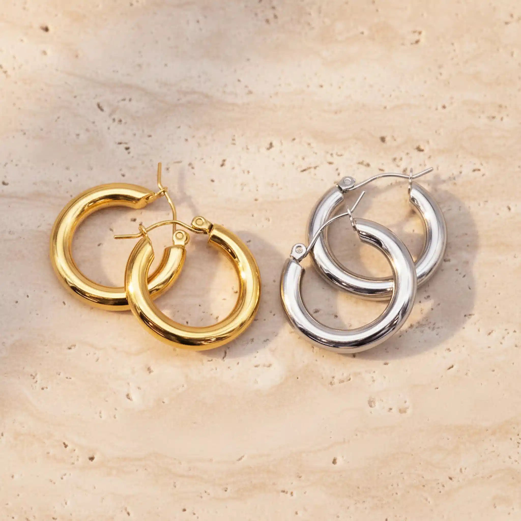 Gold and silver waterproof hoop earrings on a beige stone surface