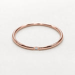A minimalist rose gold band ring with a single small clear gemstone set in the center, photographed on a clean, light gray background.