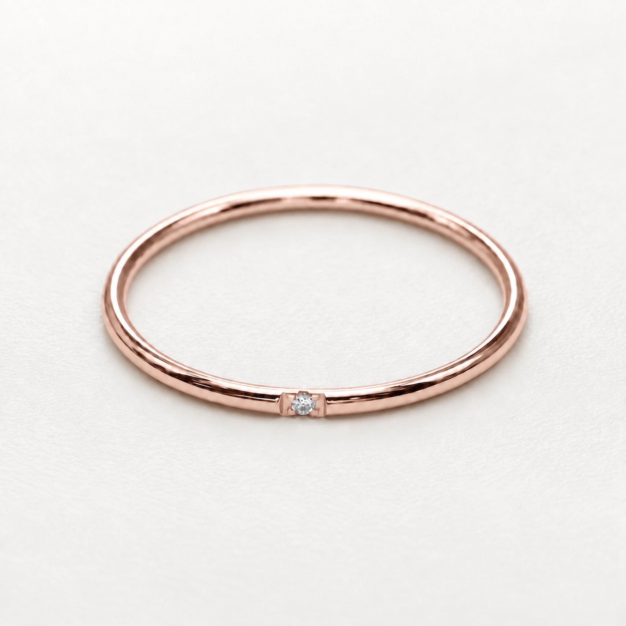 A minimalist rose gold band ring with a single small clear gemstone set in the center, photographed on a clean, light gray background.
