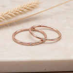 Two slim rose gold rings with small clear gemstone accents, resting on a light beige stone surface with dried wheat in the background.