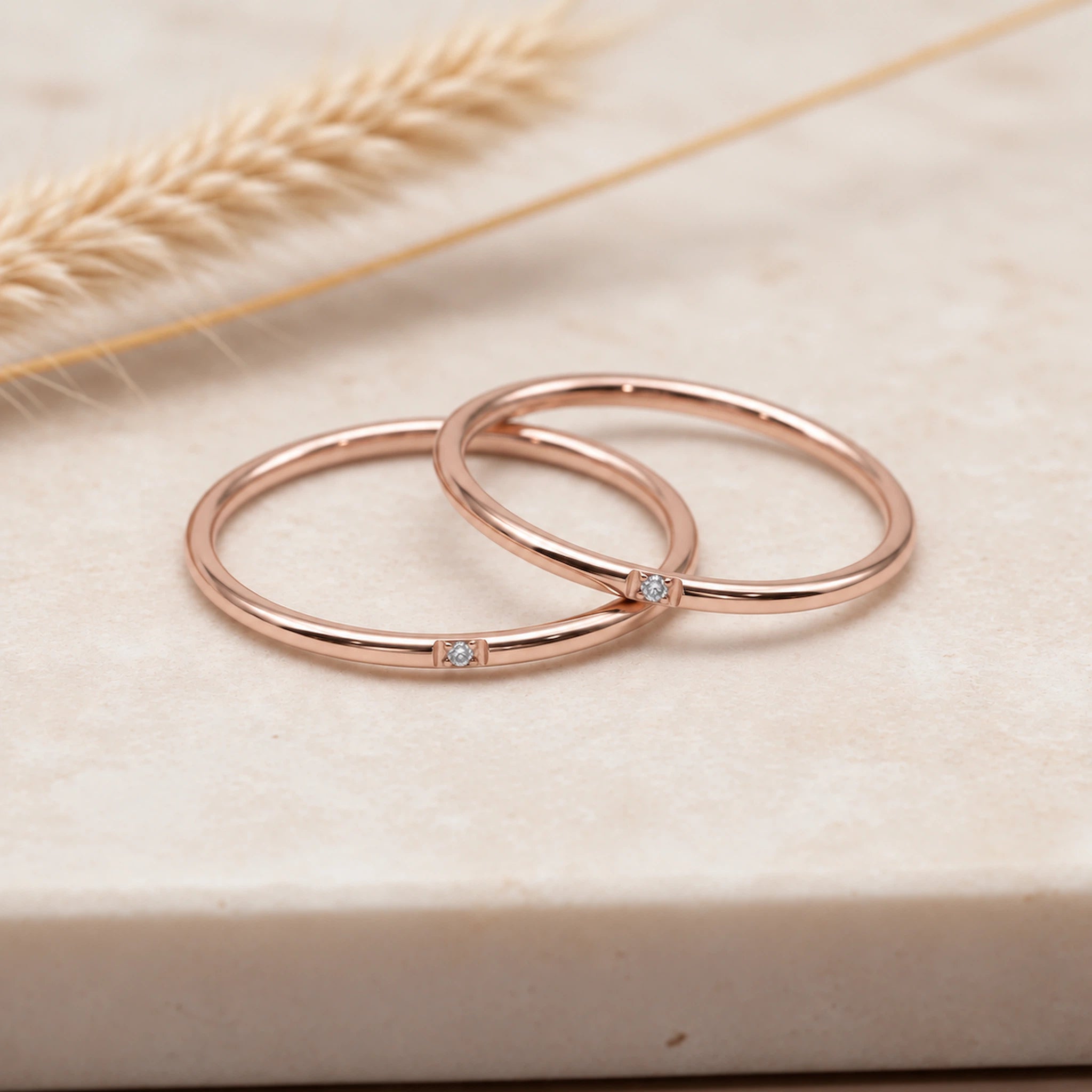 Two slim rose gold rings with small clear gemstone accents, resting on a light beige stone surface with dried wheat in the background.