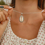 woman wearing white top with red flowers holding up a sterling silver cowrie shell necklace - women’s cowrie shell jewellery by Australian jewellery brands indie and harper