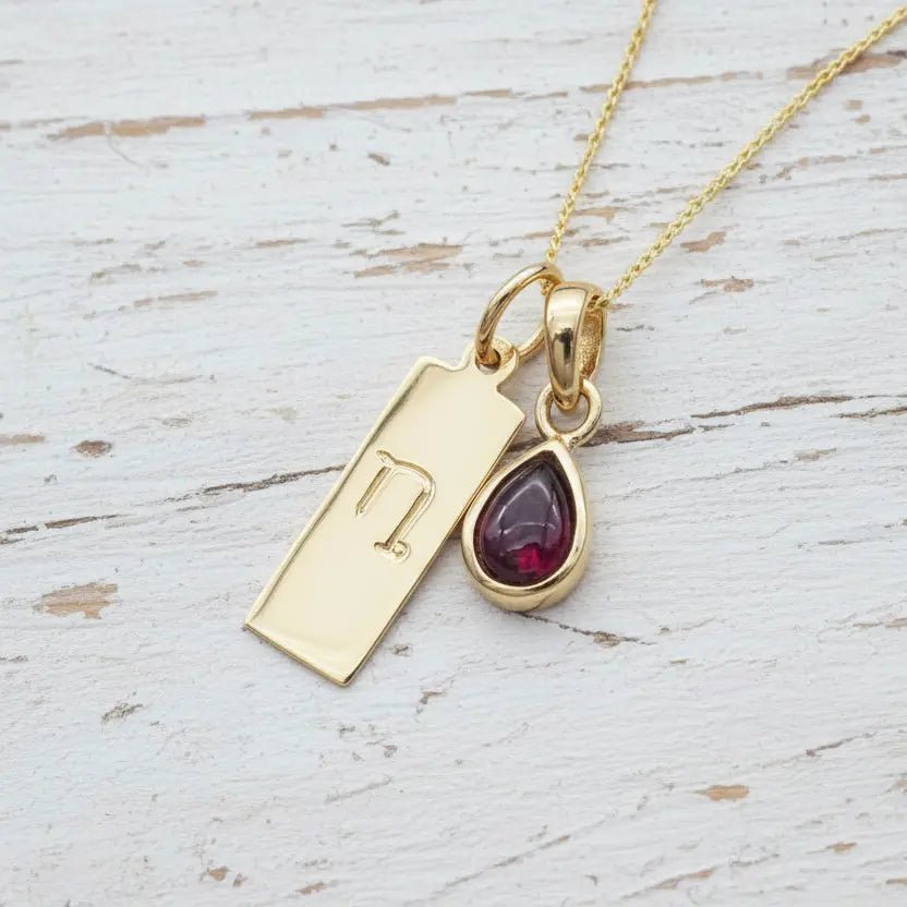 Gold necklace with Capricorn Zodiac symbol pendant and a teardrop-shaped natural garnet gemstone pendant on a light wooden surface