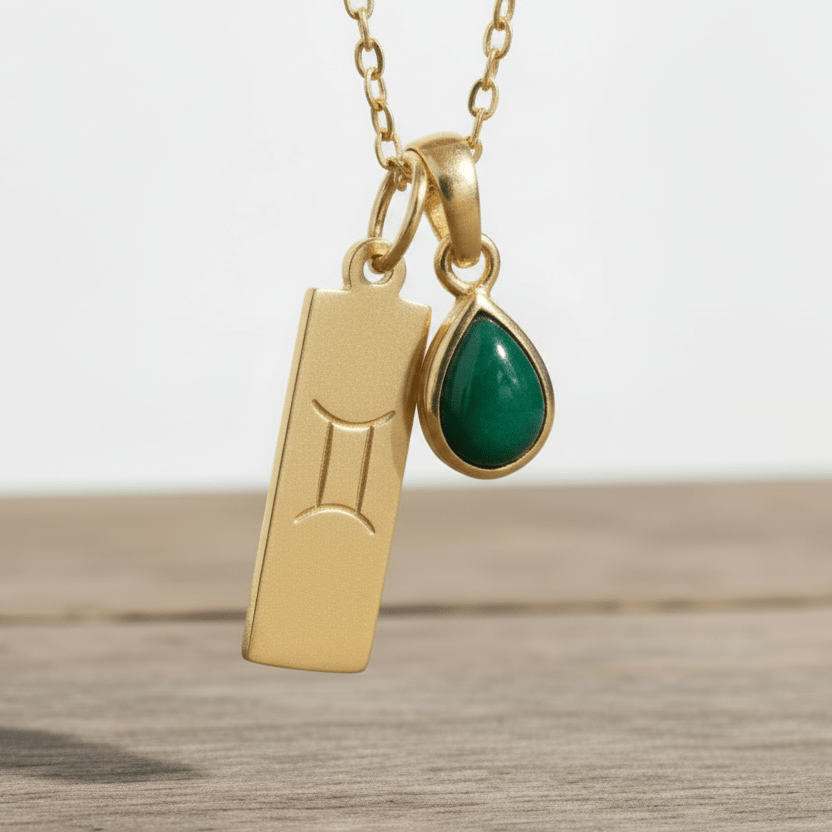 Gold necklace with a green tear drop natural emerald gemstone pendant and a gold Gemini zodiac pendant on a wooden surface