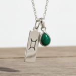 Silver necklace with a green teardrop emerald pendant and a rectangular Gemini zodiac pendant with a symbol on a wooden surface.