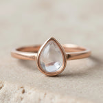 Rose gold ring with a teardrop-shaped clear herkimer quartz gemstone on a beige background
