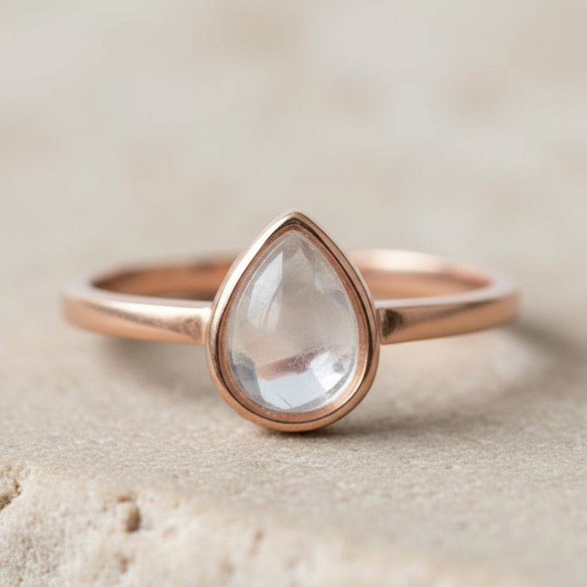 Rose gold ring with a teardrop-shaped clear herkimer quartz gemstone on a beige background