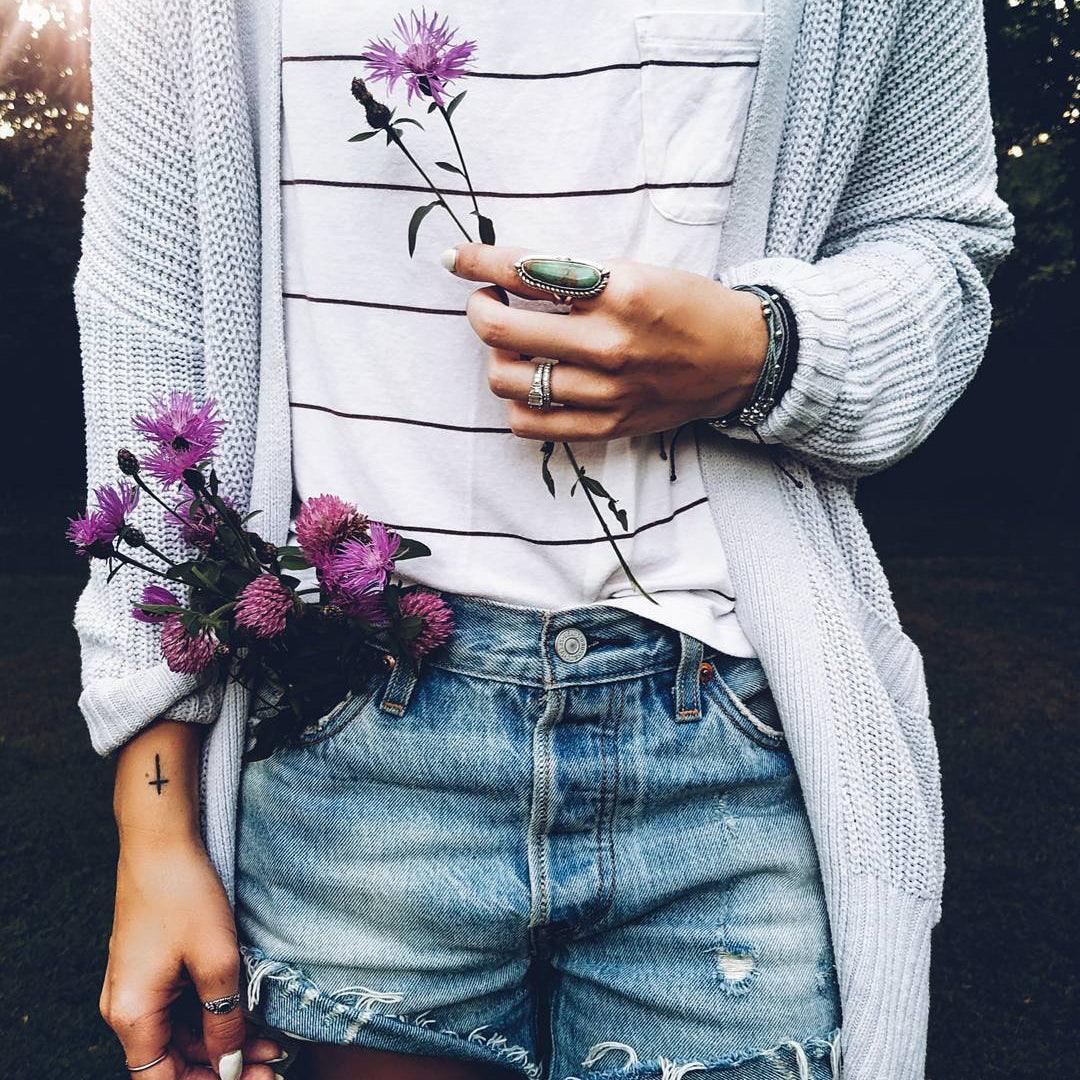 Person wearing a white sweater, striped shirt, and denim shorts holding purple flowers and wearing a silver turquoise ring