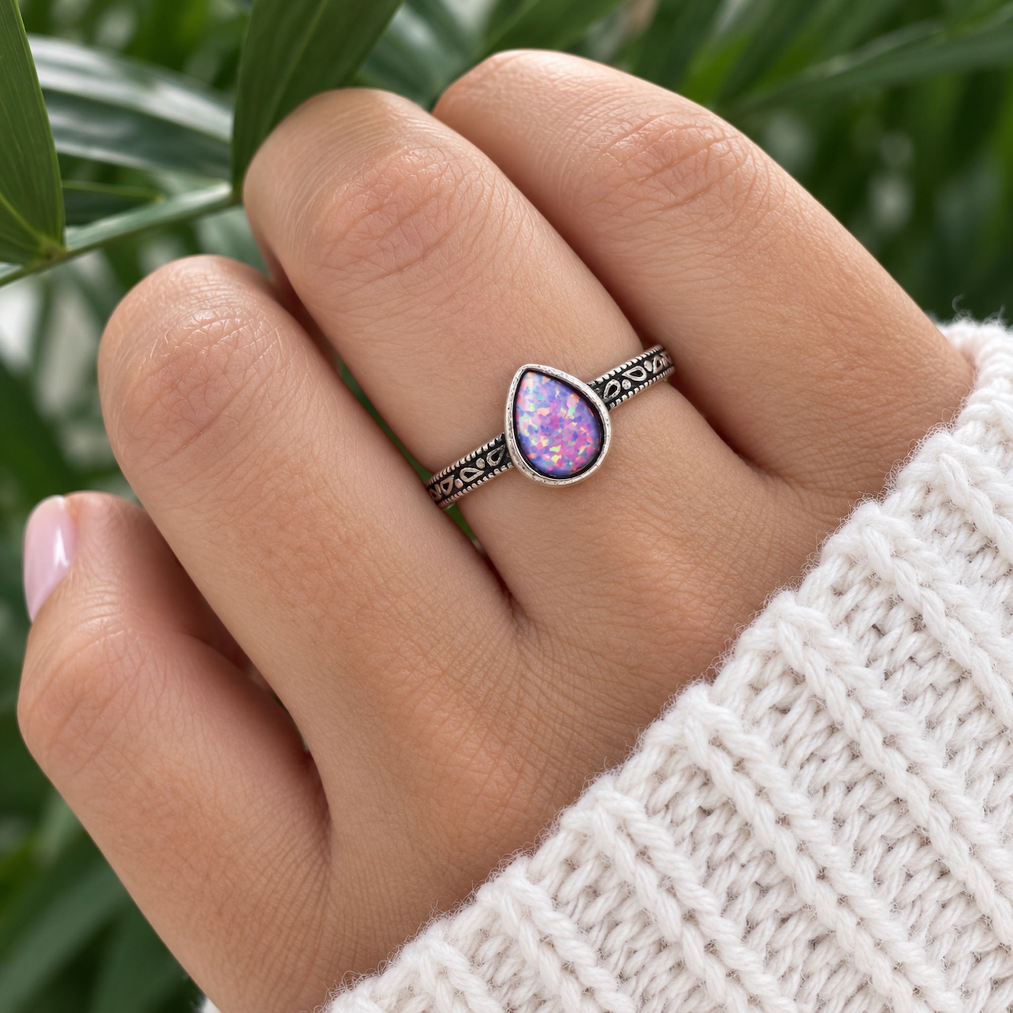 Hand wearing a sterling silver ring with a teardrop-shaped purple opal against a blurred green background